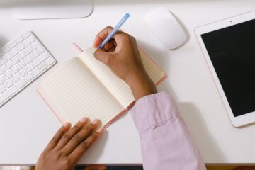 brown skin hands taking notes in planner with pen at a computer desk